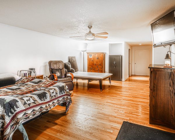Bedroom pictured from rear corner. The bed is in the foreground followed by a recliner and a mat table in the back. There is a wardrobe and a tall safe against the back wall.