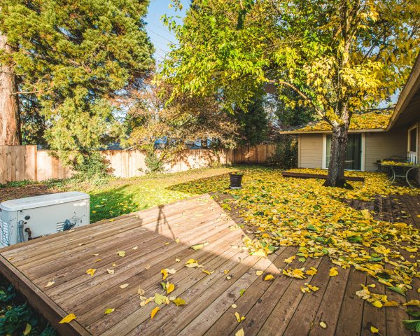 Backyard pictured from the rear corner of the deck. In the picture is the deck covered by fallen leaves from a large central tree. There is a standby generator pictured in the foreground followed by an area of grass and then the fence line.