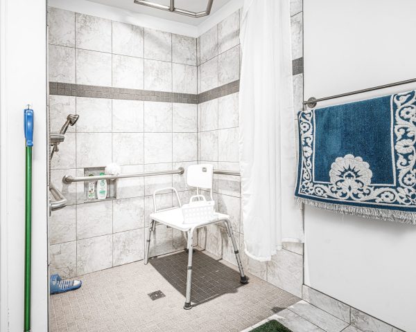 A tiled roll-in shower with grab bars, a shower chair and an adjustable height showerhead. A floor drain is in the center of the shower floor.